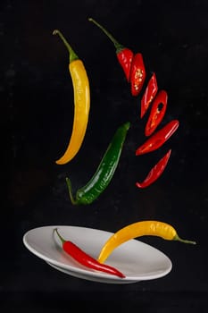 Flying red, yellow and green chili peppers on a dark background with a ceramic plate
