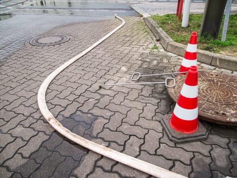 Two red and white striped warning pylons on shaped concrete blocks next to an open sewer cover with plank openings and rolled out fire hose on shaped concrete blocks in Berlin.