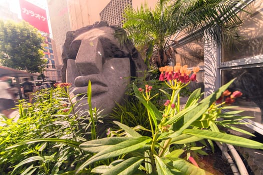 Statue of a Moai head surrounded by tropical flowers at the west exit of Shibuya Station in Tokyo, Japan. This statue was donated in 1980 by the prefecture of Nijima island of Izu archipelago to commemorate the 100 years of the transfer of the island in Tokyo Prefecture.