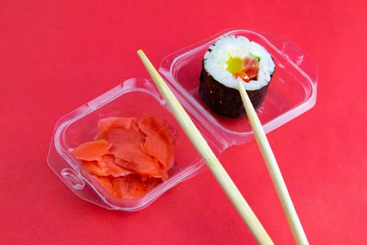 Sticks to reach for the roll, roll in the piece of packaging, next to the role of pickled ginger, everything stands on a red background. Top views