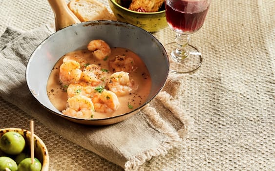 Scampi lobsters in cheese cream sauce served in old pan on rustic table with olives, bread and red wine. Traditional Spanish kitchen studio shot with copy space on linen tablecloth