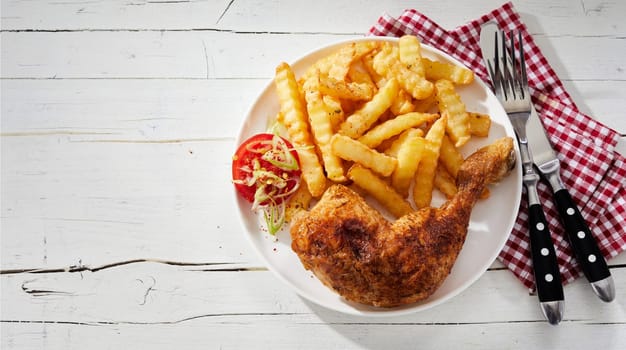 Barbecued marinated spicy seasoned chicken leg with crinkle cut potato chips, pommes frites or French fries on a plate with napkin and utensils from above with copy space