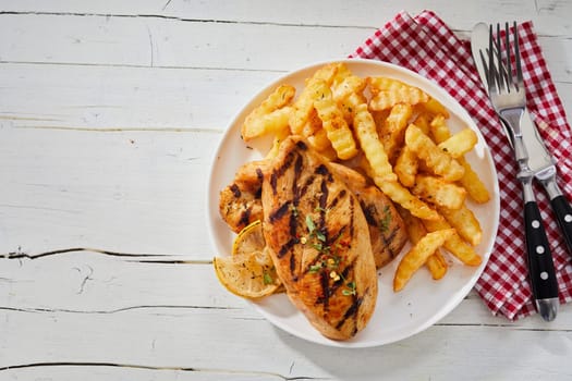 Healthy lean grilled chicken breast with crinkle cut fried potato chips or French Fries served on a plate with rustic napkin and utensils on white wood with copy space