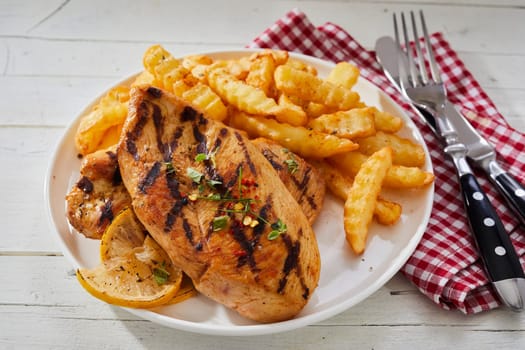 Serving of barbecued seasoned spicy chicken breast with crinkle cut potato chips on a plate with utensils and red and white napkin on rustic white table