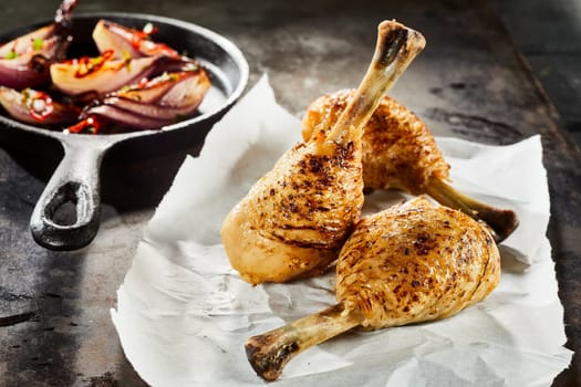 Grilled spiced chicken drumsticks with an accompaniment of roast vegetables in a skillet served on crumpled white paper