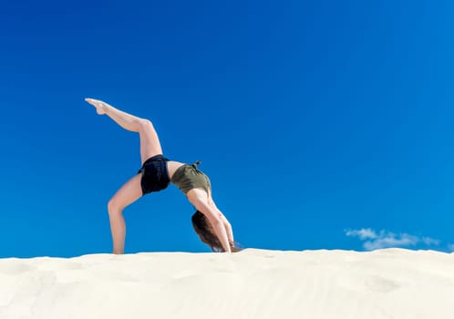 A young girl does yoga in the sand against a blue sky in summer