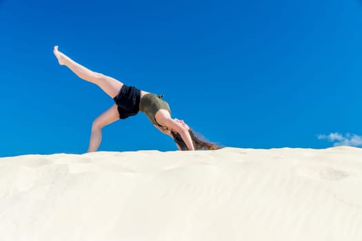 A young girl does yoga in the sand against a blue sky in summer