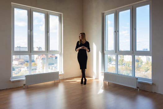 A girl in a black dress stands between two large windows in a spacious empty apartment
