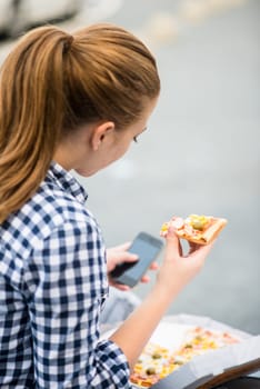 Teenager eating pizza in street and browsing internet on phone - rear view