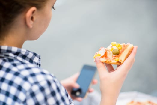 Teenager eating pizza in street and browsing internet on phone - rear view