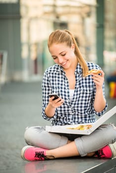 Teenager eating pizza in street and browsing internet on phone