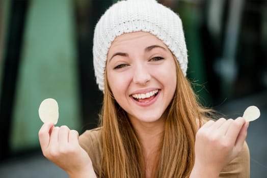Teenager girl in cap having fun with potato chips outdoor in street