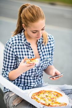 Teenager eating pizza in street and browsing internet on phone