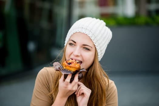 Young woman - teenager in cap eating muffin outdoor in street