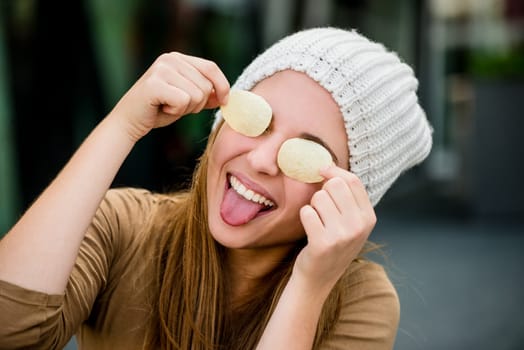 Teenager girl in cap having fun with potato chips on eyes outdoor in street