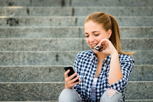 Teenager - young woman eating chocolate in street and looking in phone