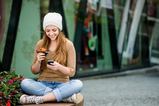 Teenager - young woman eating muffin in street and looking in phone