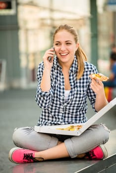 Young woman - teenage girl eating pizza in street and calling phone
