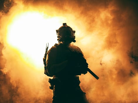 Silhouette of modern infantry soldier, elite army fighter in tactical ammunition and helmet, standing with assault service rifle in hands on background of fiery explode. Burning fire of war conflict
