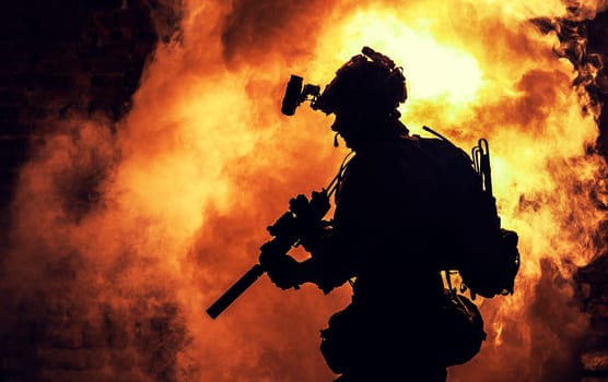 Silhouette of modern infantry soldier, elite army fighter in tactical ammunition and helmet, standing with assault service rifle in hands on background of fiery explode. Burning fire of war conflict