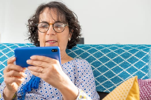 Portrait of cheerful brunette senior woman using smartphone while sitting on sofa