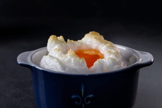 Delicate baked egg Orsini (eggs in the cloud). French breakfast in a blue cocotte against a dark background