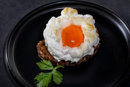 Delicate baked egg Orsini (eggs in the cloud). French breakfaston a piece of bread in a black ceramic dish