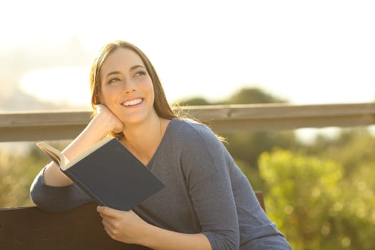 Happy woman holding a paper book thinking looking at side at sunset in a park