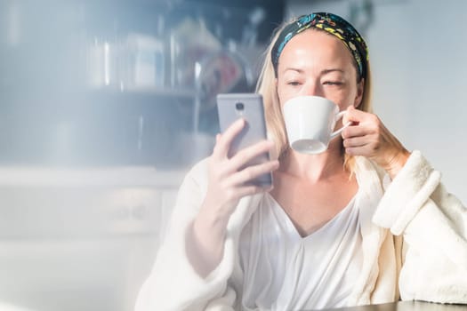Beautiful caucasian woman at home, feeling comfortable wearing white bathrobe, taking some time to herself, drinking morning coffee and reading news on mobile phone device in the morning.