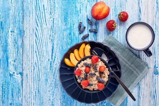 Mug with yogurt and oatmeal porridge with fresh berries and fruit in dark blue bowl on blue wooden background. Top view with copy space. Concept of healthy lifestyle, healthy food, dieting