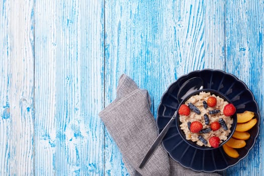 Oatmeal porridge with fresh berries and fruit in dark blue bowl on blue wooden background. Top view with copy space. Concept of healthy lifestyle, healthy food, dieting, vegetarian food