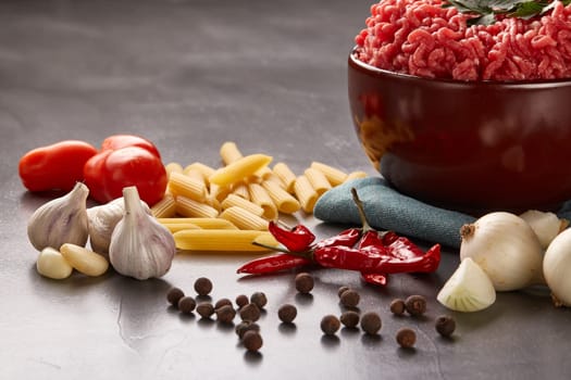 Still life with fresh forcemeat from beef in a bowl, vegetables, pasta and spices on a dark background.