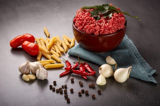 Still life with fresh forcemeat from beef in a bowl, vegetables, pasta and spices on a dark background.