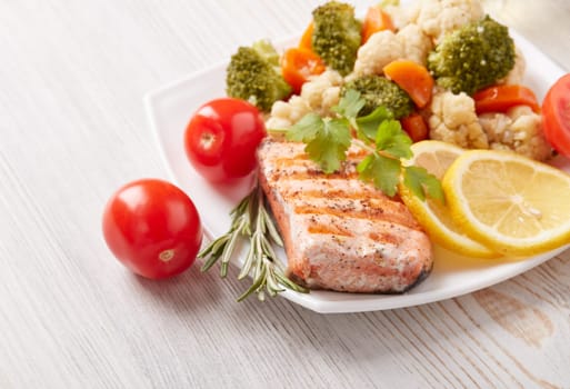Grilled salmon fillet  garnished with vegetables, lemon, tomato, herbs lying on plate on wooden background. Top view. Healthy food. Keto diet.