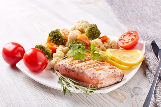 Grilled salmon fillet  garnished with vegetables, lemon, tomato, herbs lying on plate on wooden background. Healthy food. Keto diet.