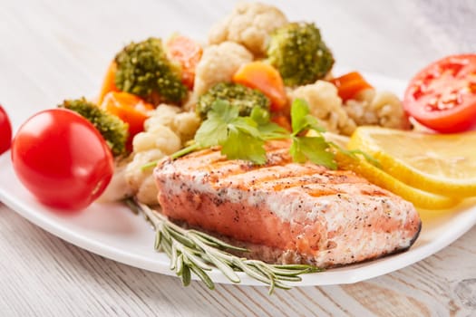 Grilled salmon fillet  garnished with vegetables, lemon, tomato, herbs lying on plate on wooden background. Healthy food. Keto diet.