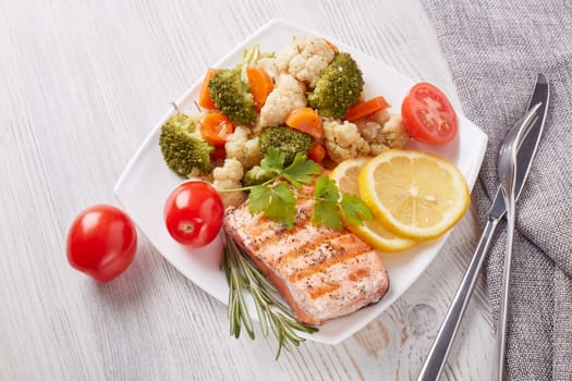 Grilled salmon fillet  garnished with vegetables, lemon, tomato, herbs lying on plate on wooden background. Top view.Healthy food. Keto diet.