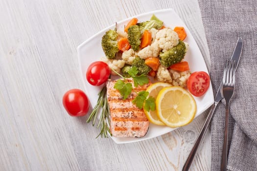 Grilled salmon fillet  garnished with vegetables, lemon, tomato, herbs lying on plate on wooden background. Top view with copy space.Healthy food. Keto diet.