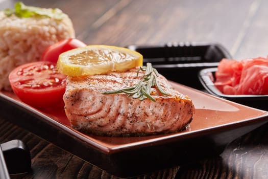 Grilled salmon fillet with rice, pickled ginger, soy sauce, tomato, lemon and herbs on dark wooden background.  Healthy food.