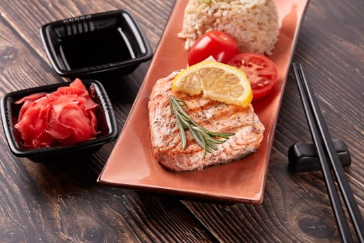 Grilled salmon fillet with rice, pickled ginger, soy sauce, tomato, lemon and herbs on dark wooden background. Top view. Healthy food.