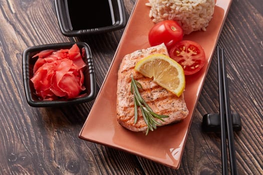 Grilled salmon fillet with rice, pickled ginger, soy sauce, tomato, lemon and herbs on dark wooden background. Top view. Healthy food.