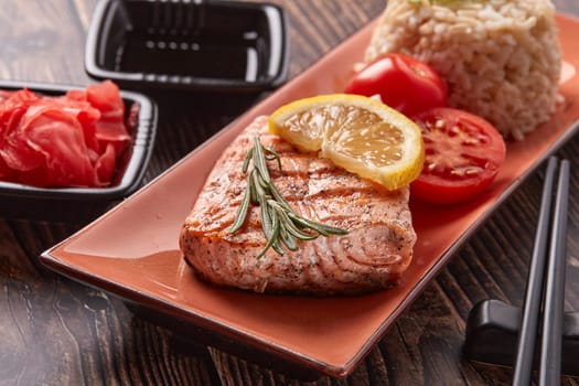 Grilled salmon fillet with rice, pickled ginger, soy sauce, tomato, lemon and herbs on dark wooden background.  Healthy food.
