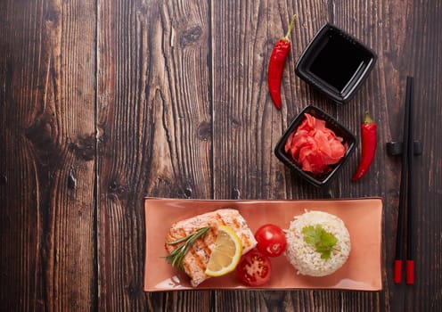 Grilled salmon fillet with rice, pickled ginger, soy sauce, tomato, lemon and herbs on dark wooden background. Top view with copy space. Healthy food.