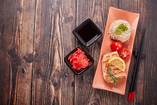 Grilled salmon fillet with rice, pickled ginger, soy sauce, tomato, lemon and herbs on dark wooden background. Top view with copy space. Healthy food.