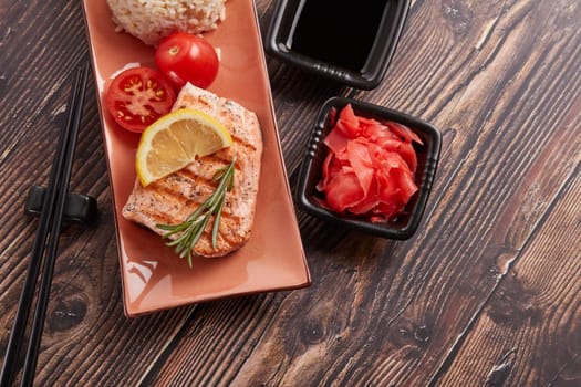 Grilled salmon fillet with rice, pickled ginger, soy sauce, tomato, lemon and herbs on dark wooden background. Top view. Healthy food.