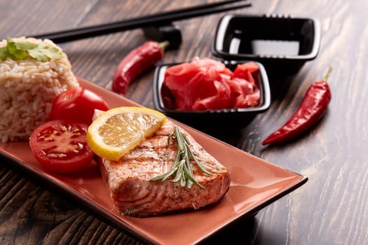 Grilled salmon fillet with rice, pickled ginger, soy sauce, tomato, lemon and herbs on dark wooden background.  Healthy food.