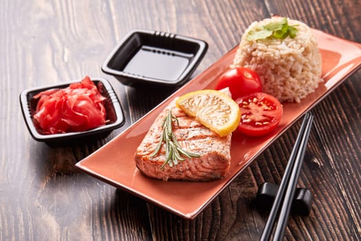 Grilled salmon fillet with rice, pickled ginger, soy sauce, tomato, lemon and herbs on dark wooden background.  Healthy food.