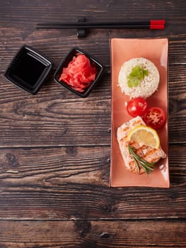 Grilled salmon fillet with rice, pickled ginger, soy sauce, tomato, lemon and herbs on dark wooden background. Top view with copy space. Healthy food.