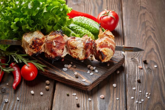 Shashlik from chicken with bacon. Grilled chicken (turkey) fillet on skewer and fresh vegetables with spices on stone cutting board on wooden background