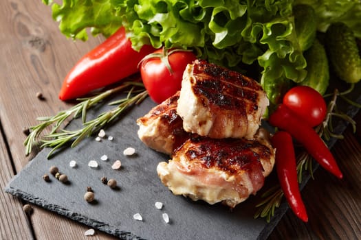Bacon-wrapped grilled chicken (turkey)  fillet with fresh vegetables and spices on stone cutting board on wooden background